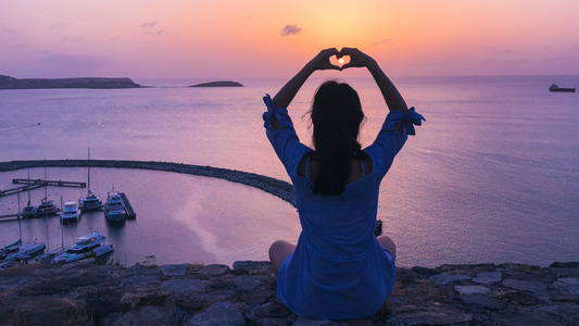 Woman sitting with her arms above her head with her hands in a heart shape over a sunset bay